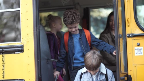 Cute diverse elementary age schoolkids saying bye to smiling mixed race woman driver while getting off school bus. Preadolescent friendly schoolchildren taking turns leaving bus arriving at school