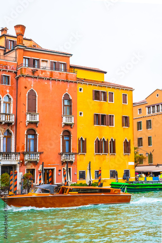 Luxury wooden water taxi boat/ yacht sailing Grand Canal water/ waterway by colorful gothic Venetian architecture hotel, other buildings and striped mooring poles. Venice city, Italy in rainy November