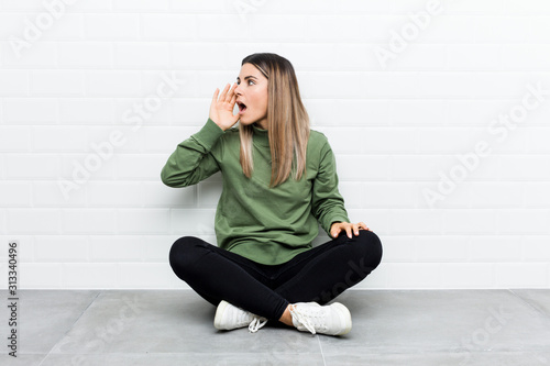 Young caucasian woman sitting on the floor shouting and holding palm near opened mouth.