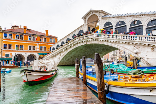 Rainy November day/ season in Venice, Italy. People/ tourists walk/ stand with umbrellas on Rialto Bridge on Grand Canal. Colorful boats docked and sailing on water below. Famous landmark attraction.