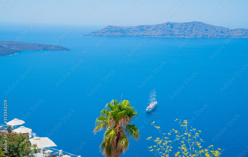Four-mast cruise ship in harbor below from hill in Fira with palm tree and yellow flowers and sun-umbrellas on balcony.