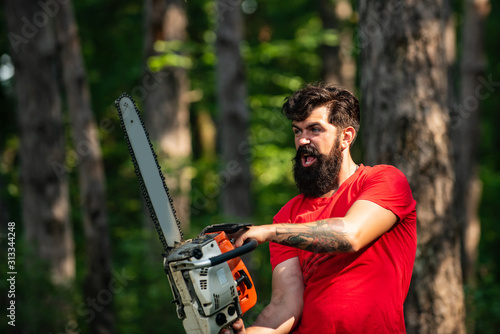 Lumberjack on serious face carries chainsaw. Woodcutter with chainsaw on sawmill. A handsome young man with a beard carries a tree. Harvest of timber. photo