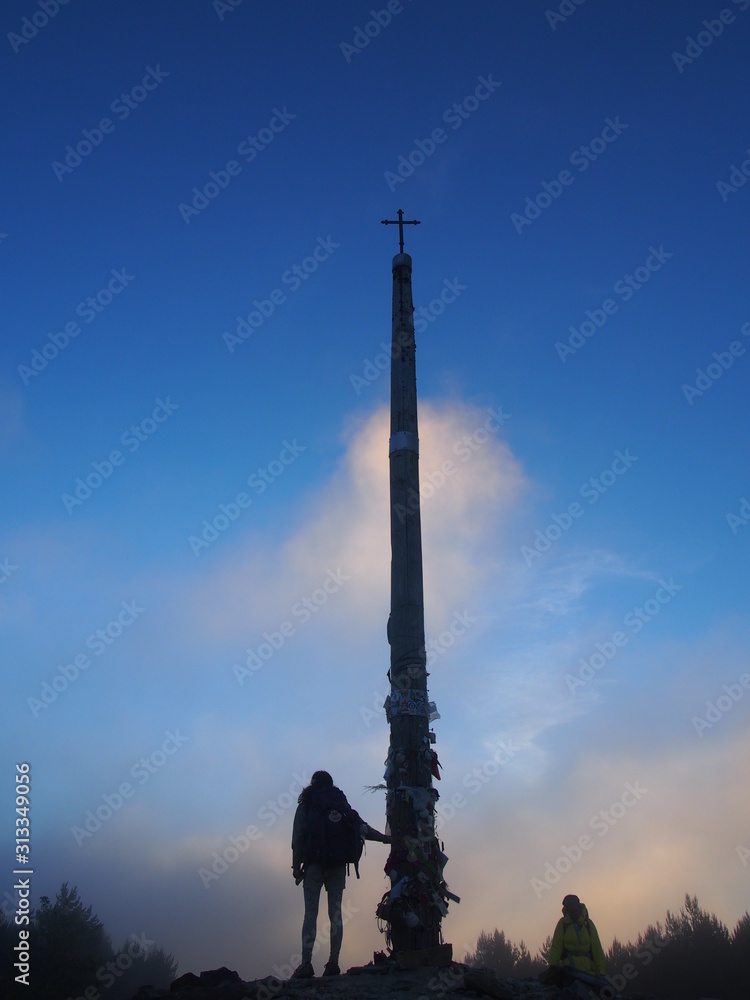 Fototapeta premium Pilgrim and beautiful blue sky with Cruz de Ferro (Cross of Iron) in the early morning, Camino de Santiago, Way of St. James, Foncebadon to Ponferrada, French way, Spain