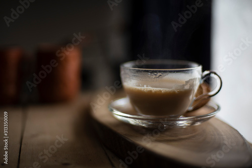 A transparent glass cup and soccer filled up with infused tea/coffee is kept in a wooden tray/table in black background in the winter morning. Indian beverages and food photography.
