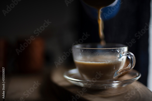 Hot infused tea coffee being poured in a transparent glass cup and soccer kept in a wooden tray table from a kettle in black background in the winter morning. Indian beverages and food photography.