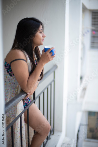An young and attractive Indian brunette woman in white sleeping wear standing in a balcony with a tea/coffee mug in the morning in white background. Indian lifestyle.