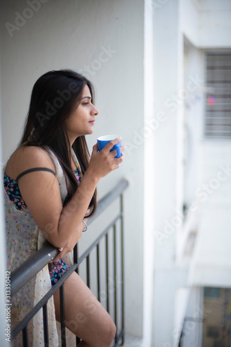 An young and attractive Indian brunette woman in white sleeping wear standing in a balcony with a tea/coffee mug in the morning in white background. Indian lifestyle.