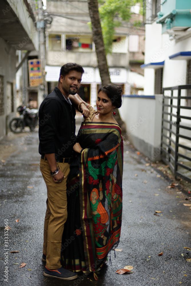 A brunette Indian Bengali romantic couple in traditional wear interacting between themselves in the drenched street in the morning of Durga Puja festival in urban background. Indian lifestyle.