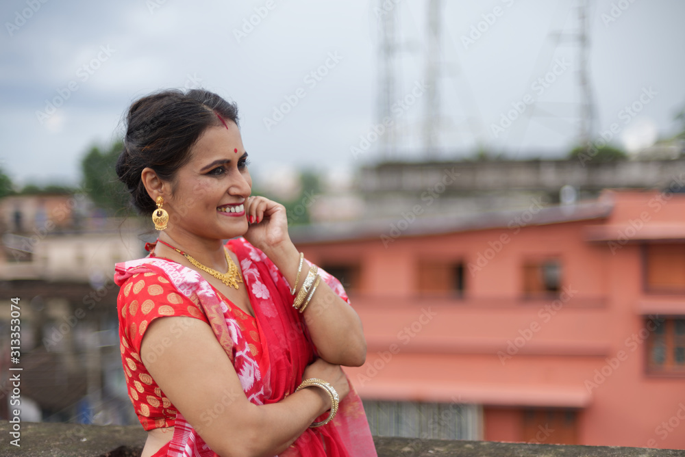 Portrait of a beautiful smiling brunette Indian Bengali woman in traditional red sari standing on the roof top under blue sky in afternoon of Durga Puja festival in urban background. Indian lifestyle.