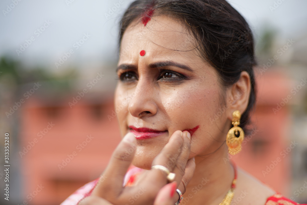 Portrait of beautiful smiling brunette Indian Bengali woman in traditional red sari having vermillion on cheek on the roof top in afternoon of Durga Puja festival in urban background.Indian lifestyle. Portrait of beautiful smiling brunette Indian Bengali woman in traditional red sari having vermillion on cheek on the roof top in afternoon of Durga Puja festival in urban background.Indian lifestyle.