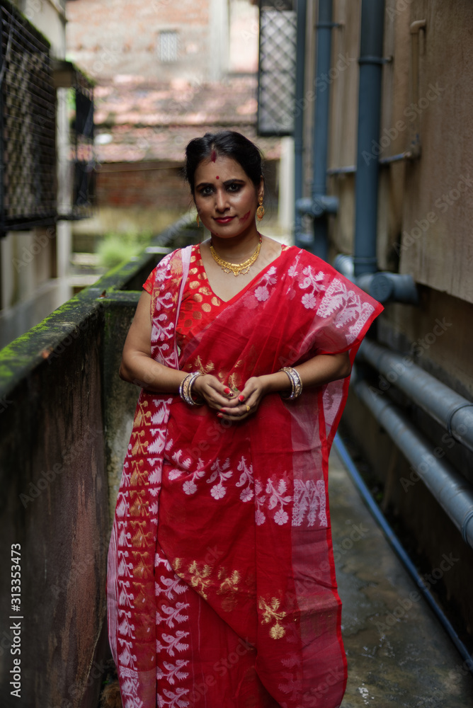 Portrait of a beautiful smiling brunette Indian Bengali woman in traditional red sari standing in a narrow lane in the morning of Durga Puja festival in urban background. Indian lifestyle.