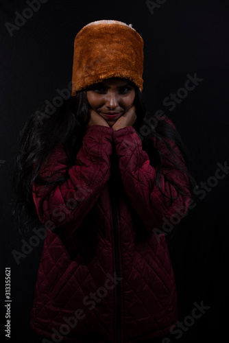Portrait of an young and attractive dark skinned Indian woman wearing woolen cap and jacket feeling cold in a winter night in dark background. Winter and Christmas