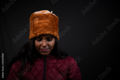 Portrait of an young and attractive dark skinned Indian woman wearing woolen cap and jacket feeling cold in a winter night in dark background. Winter and Christmas