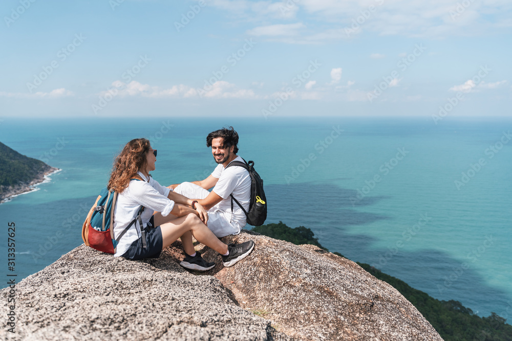 Happy beautiful young couple of travelers man and woman on top of a mountain with ocean view. Romantic travels, honeymoon,