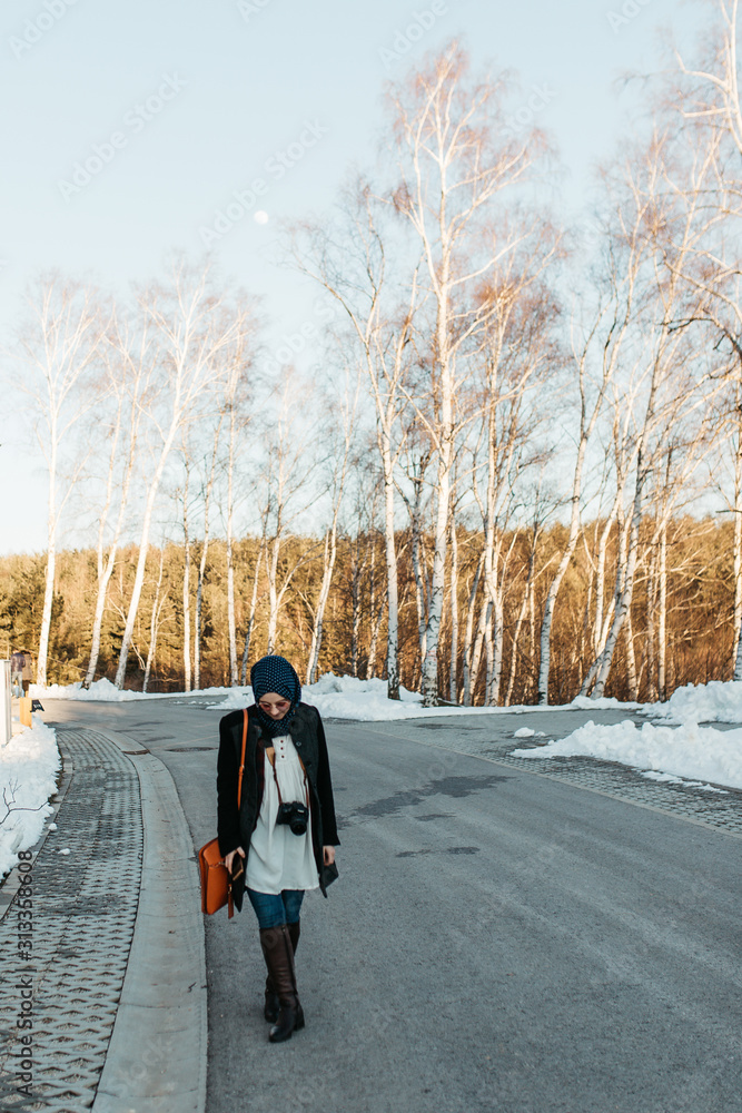 portrait of young European Muslim women with hijab standing on the edge of the mountain. She is holding hat in one hand and waving with the other. She is also holding camera and taking pictures.