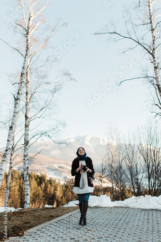 portrait of young European Muslim women with hijab standing on the edge of the mountain. She is holding hat in one hand and waving with the other. She is also holding camera and taking pictures.