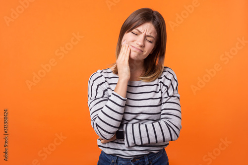 Portrait of unhappy sick young woman with brown hair in long sleeve striped shirt standing, touching cheek, suffering acute toothache, need dentist indoor studio shot isolated on orange background