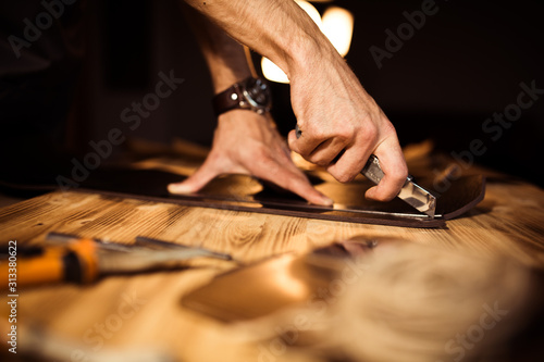 Working process of the leather belt in the leather workshop. Man holding crafting tool and working. Tanner in old tannery. Wooden table background. Close up man arm.