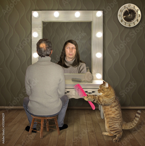 The balding man in a sweater is looking in the mirror at home. He sees himself with long hair in his reflection. His cat gives him a comb. photo