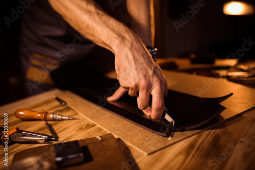 Working process of the leather belt in the leather workshop. Man holding crafting tool and working. Tanner in old tannery. Wooden table background.