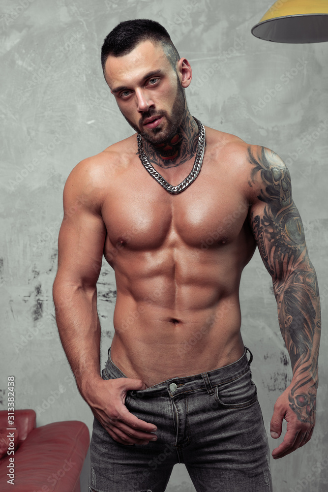 Fashion portrait of Sexy naked male model with tattoo and a black beard  standing in hot pose. Loft room interior with grey concrete wall.  Professional Studio image. Stock-bilde | Adobe Stock