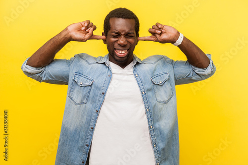 Don't want to hear. Portrait of frustrated irritated man in denim casual shirt covering ears with annoyed expression, ignoring conflict or pressure. indoor studio shot isolated on yellow background