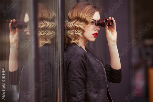 Beautiful brunette sexy spy agent (killer or police) woman in leather jacket and red luxury dress with a binocular in her hand stalking someone, to catch him in european city