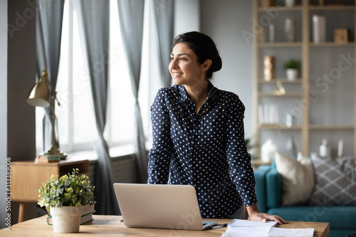 Dreamy indian woman looking away dreaming at home with laptop