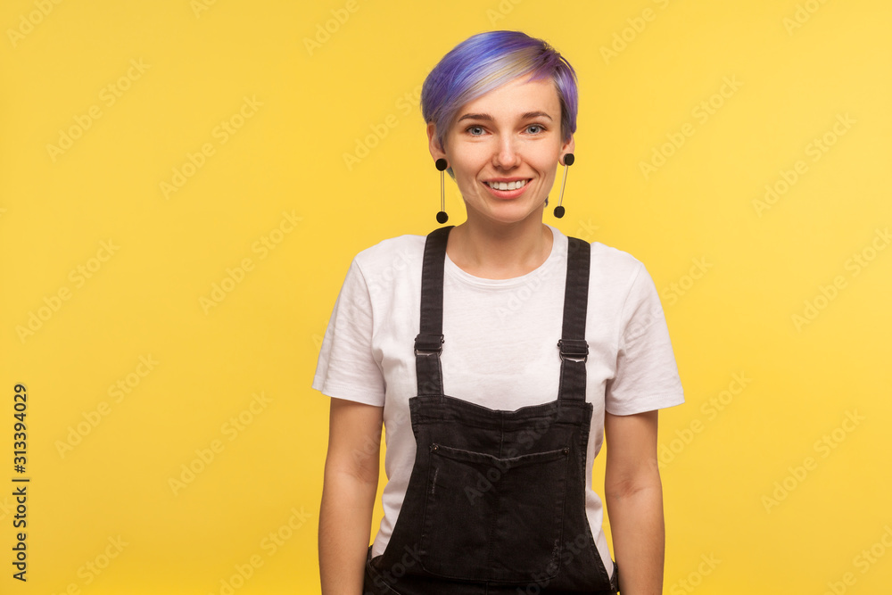 Portrait of happy trendy hipster woman with violet short hair in overalls looking at camera with toothy smile, positive mood, stylish fashionable appearance. isolated on yellow background, studio shot Portrait of happy trendy hipster woman with violet short hair in overalls looking at camera with toothy smile, positive mood, stylish fashionable appearance. isolated on yellow background, studio shot