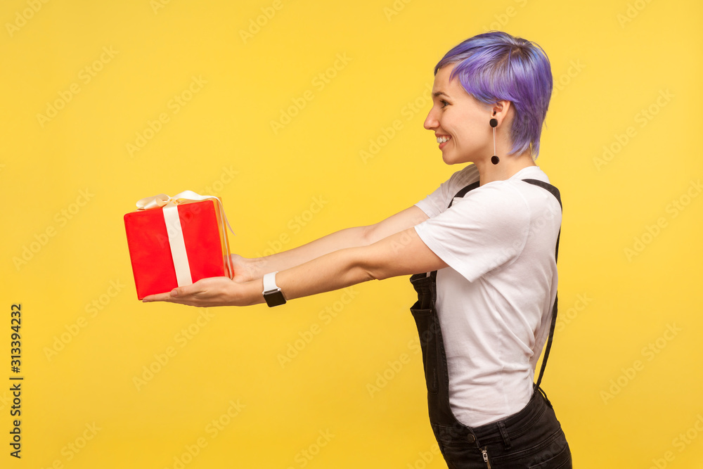 Take present! Side view portrait of happy trendy fashionable girl with violet short hair in overalls giving present box and smiling, holiday charity. indoor studio shot isolated on yellow background Take present! Side view portrait of happy trendy fashionable girl with violet short hair in overalls giving present box and smiling, holiday charity. indoor studio shot isolated on yellow background
