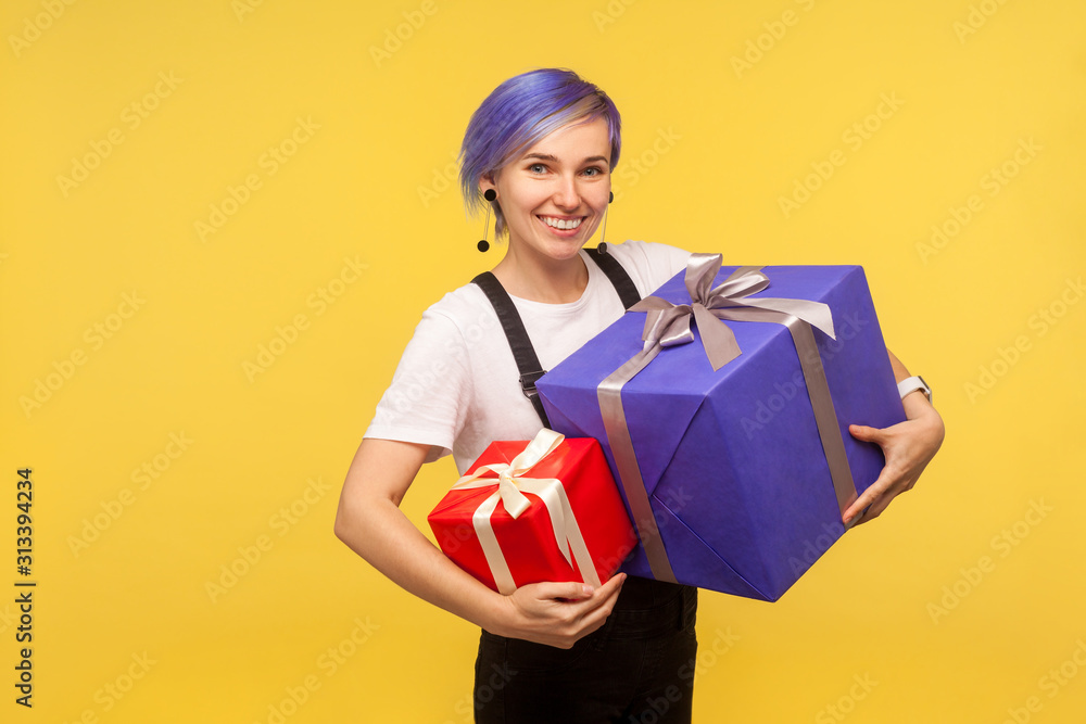 Portrait of admired joyful hipster girl with violet short hair in overalls holding two gifts, big and small holiday present boxes, looking at camera with toothy smile. yellow background, studio shot Portrait of admired joyful hipster girl with violet short hair in overalls holding two gifts, big and small holiday present boxes, looking at camera with toothy smile. yellow background, studio shot
