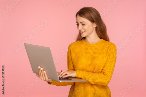 Portrait of happy positive ginger girl in sweater holding laptop, typing on keyboard, surfing internet, student or freelancer working using computer. indoor studio shot isolated on pink background