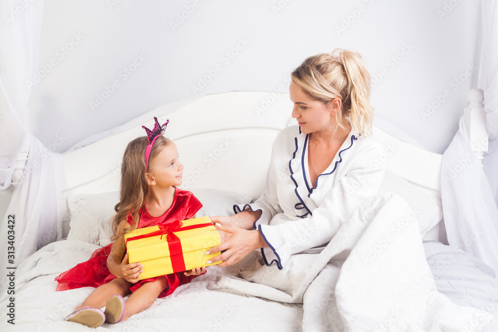 Young woman giving present to little cute girl in a bright dress and crown on head, sitting on bed, mother wishing daughter happy birthday, greeting child on holiday morning, congratulation in bedroom Young woman giving present to little cute girl in a bright dress and crown on head, sitting on bed, mother wishing daughter happy birthday, greeting child on holiday morning, congratulation in bedroom