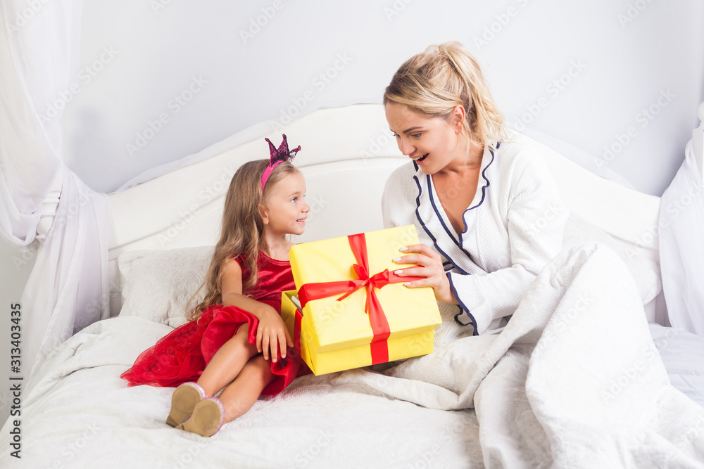 Young mother opening gift box with surprised amazed face, sitting with cute little daughter on bed, child wishing woman happy birthday, greeting mom on holiday morning, congratulation in bedroom Young mother opening gift box with surprised amazed face, sitting with cute little daughter on bed, child wishing woman happy birthday, greeting mom on holiday morning, congratulation in bedroom