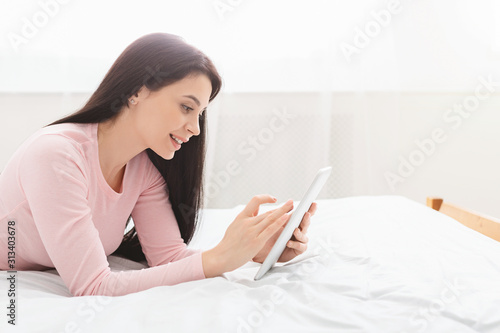 Cheerful girl browsing on digital tablet, resting in bed