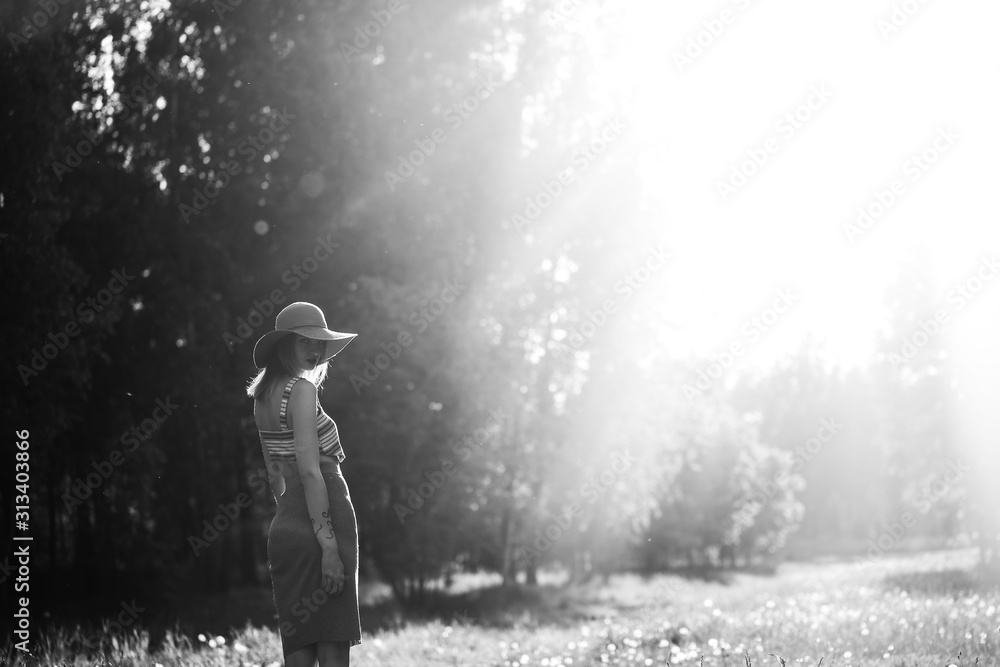 Obraz premium Silhouette of girl walking in park outdoor. Sunny summer lifestyle concept. Woman in dress and hat in field with dandelions. Light effect for text. Copyspace for design. BW photo