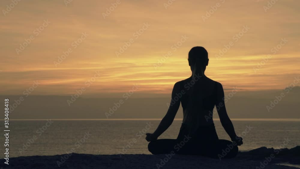 Silhouette of a meditating young woman sitting in lotus position on a rock by the sea against the background of dawn Silhouette of a meditating young woman sitting in lotus position on a rock by the sea against the background of dawn