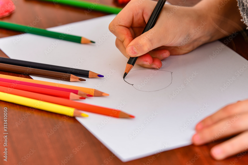 A girl draws a heart with colored pencils on a white sheet among children's toys. A girl draws a heart with colored pencils on a white sheet among children's toys.