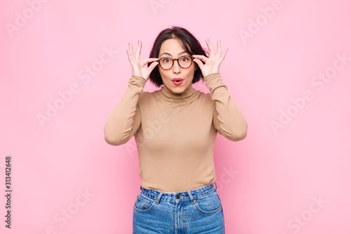 young pretty woman feeling shocked, amazed and surprised, holding glasses with astonished, disbelieving look against pink wall photo