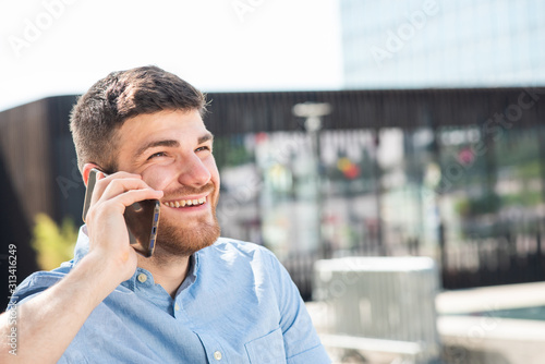 Side of happy young man with beard talking on cellphone outside city