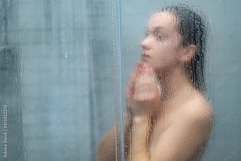 Young naked woman showering and bathing in morning shower under refreshing water jet through the bath screen with little drops