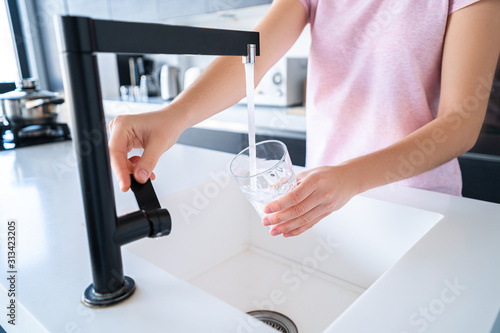 Woman pours fresh filtered purified water for drinking from a tap into a glass at kitchen at home. Healthy lifestyle