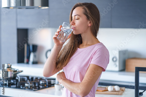Happy attractive joyful brunette woman drinking fresh clean filtered purified water at kitchen at home. Healthy lifestyle and quench thirst