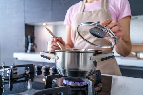 Fototapeta Woman housewife in apron using steel metal saucepan for preparing dinner in the kitchen at home