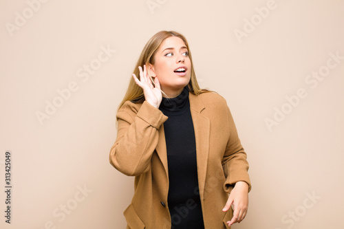 young blonde woman smiling, looking curiously to the side, trying to listen to gossip or overhearing a secret against flat wall photo