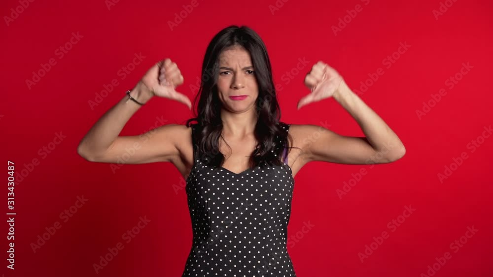 Young pretty woman standing on red studio background expressing discontent and showing thumb down gesture at camera. Portrait of girl with sign of dislike. Young pretty woman standing on red studio background expressing discontent and showing thumb down gesture at camera. Portrait of girl with sign of dislike.