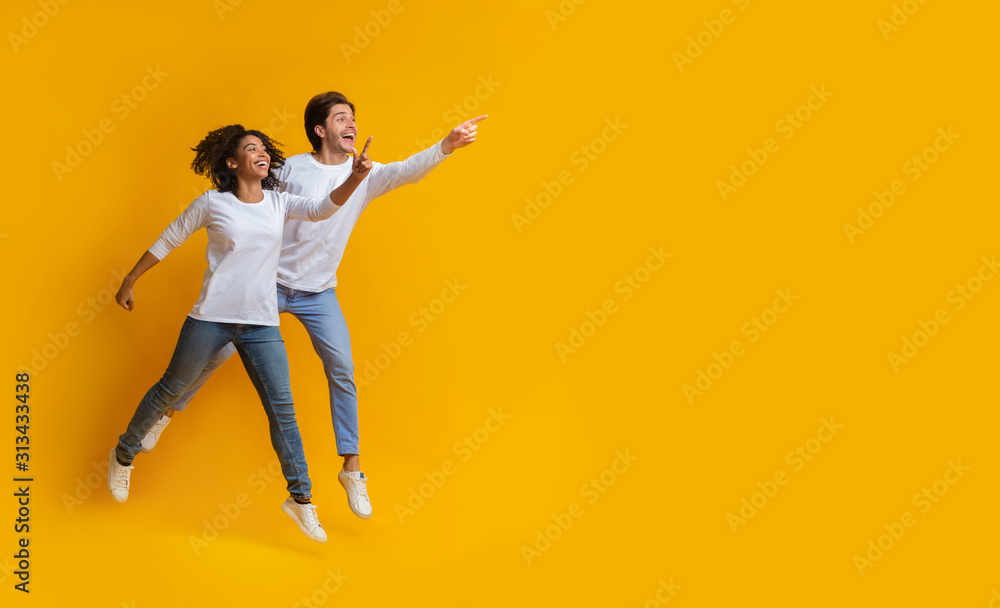 Multiracial couple pointing at copy space and jumping over yellow background Multiracial couple pointing at copy space and jumping over yellow background