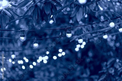 Vintage Edison incandescent bulbs garland in the street with sky, trees crowns and buildings on the background. Toned in classic blue