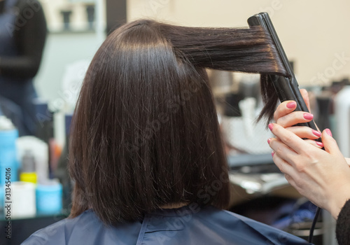 The hairdresser does aligns the hair with hair iron to a young girl, brunette in a beauty salon. Professional Hair Care