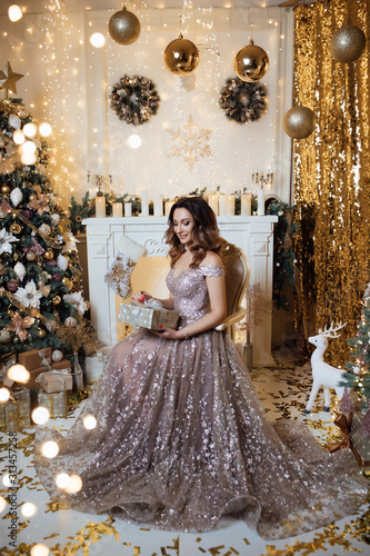 Lovely girl in dress in the New Year’s interior. Woman at home at Christmas time in a living room in a celebrate style near a New Year tree. Christmas holidays. Woman with christmas box gift.  © Tetiana Moish